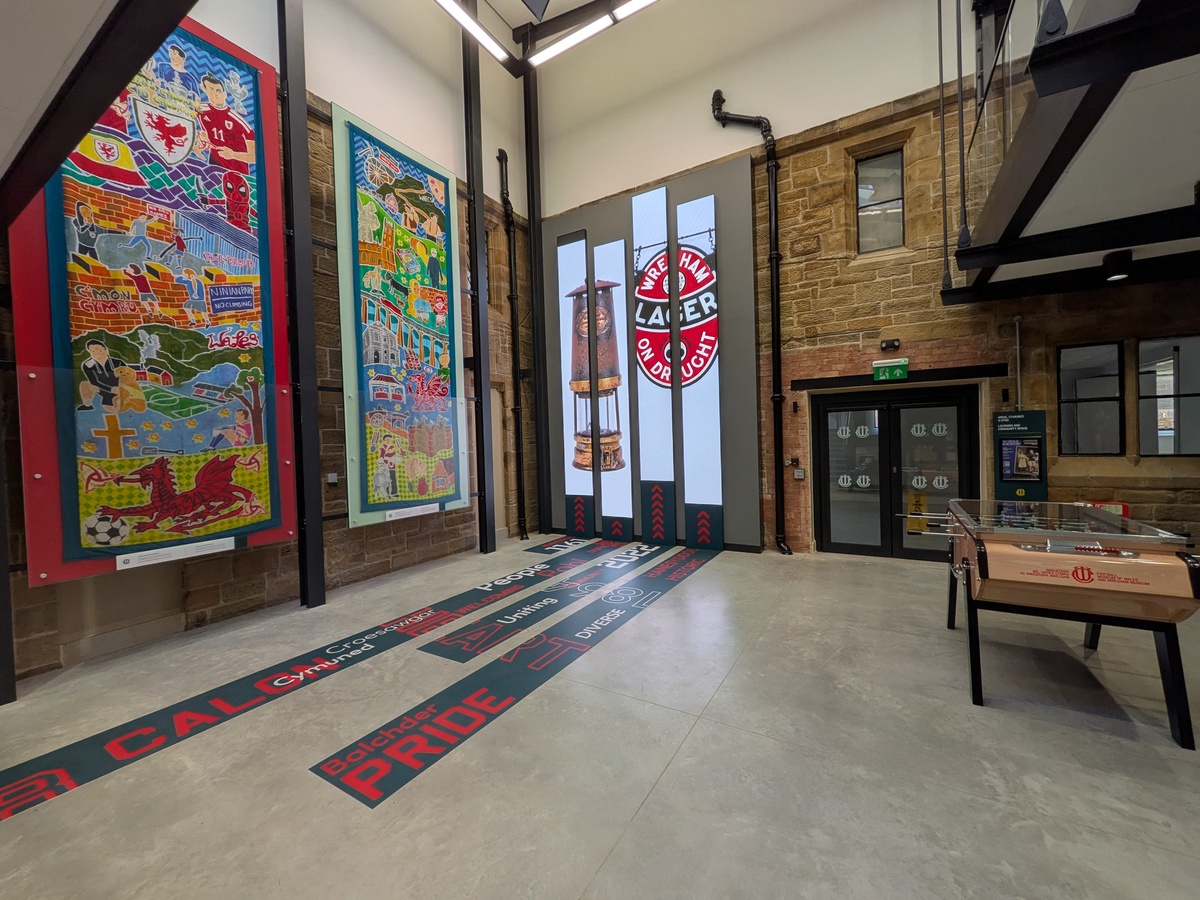 Exhibition hall with painted banners on a stone wall, a miner's lamp display, floor lettering reading PRIDE and a table football game