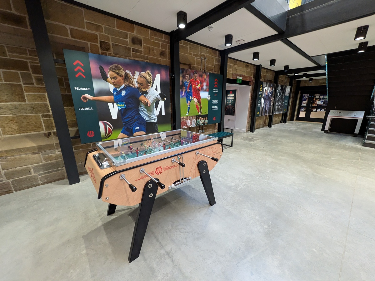 Table football in an exhibition space with stone walls, black beams and bilingual football display boards reading Pel-Droed