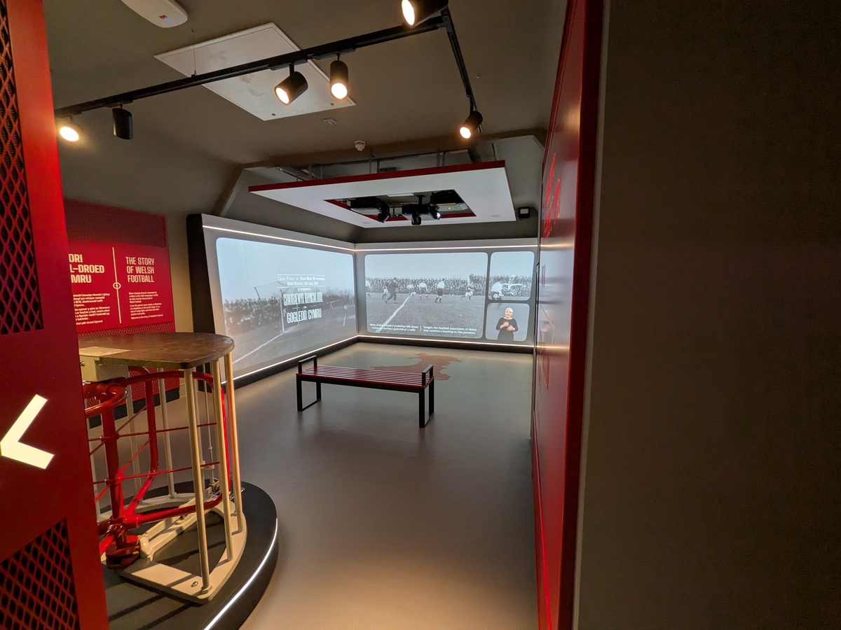 Museum gallery with a curved screen showing archive football footage, a bench, and a red turnstile on a plinth